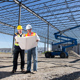 Two construction employees with hard hats, vests and blueprint standing under metal construction